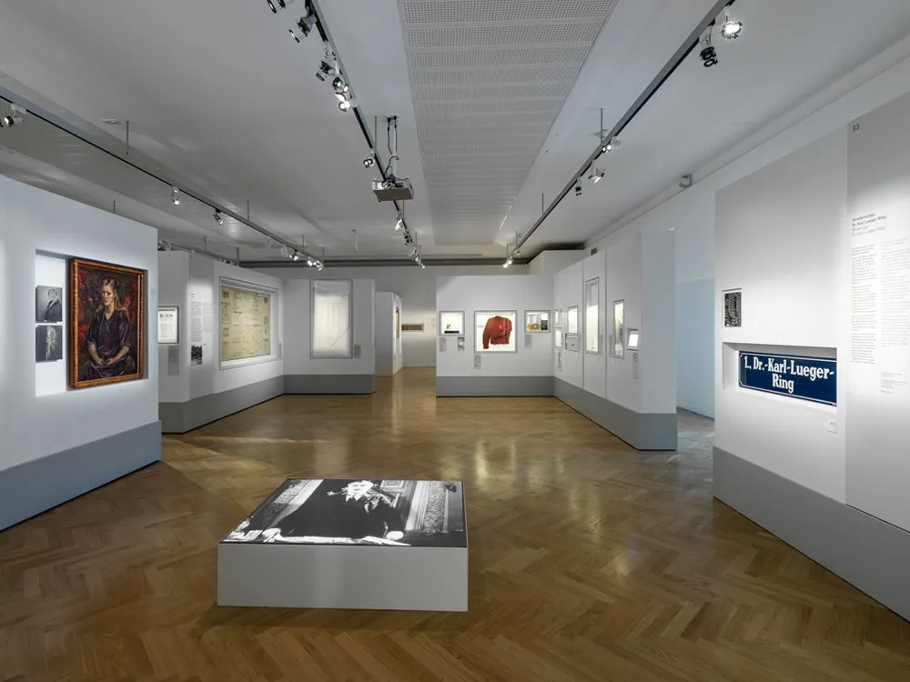 Ausstellungsraum mit einem Holzboden, weißen Wänden, Vitrinen mit Gemälden, Dokumenten und einem roten Kleid, sowie einem Tisch mit einem Foto im Zentrum