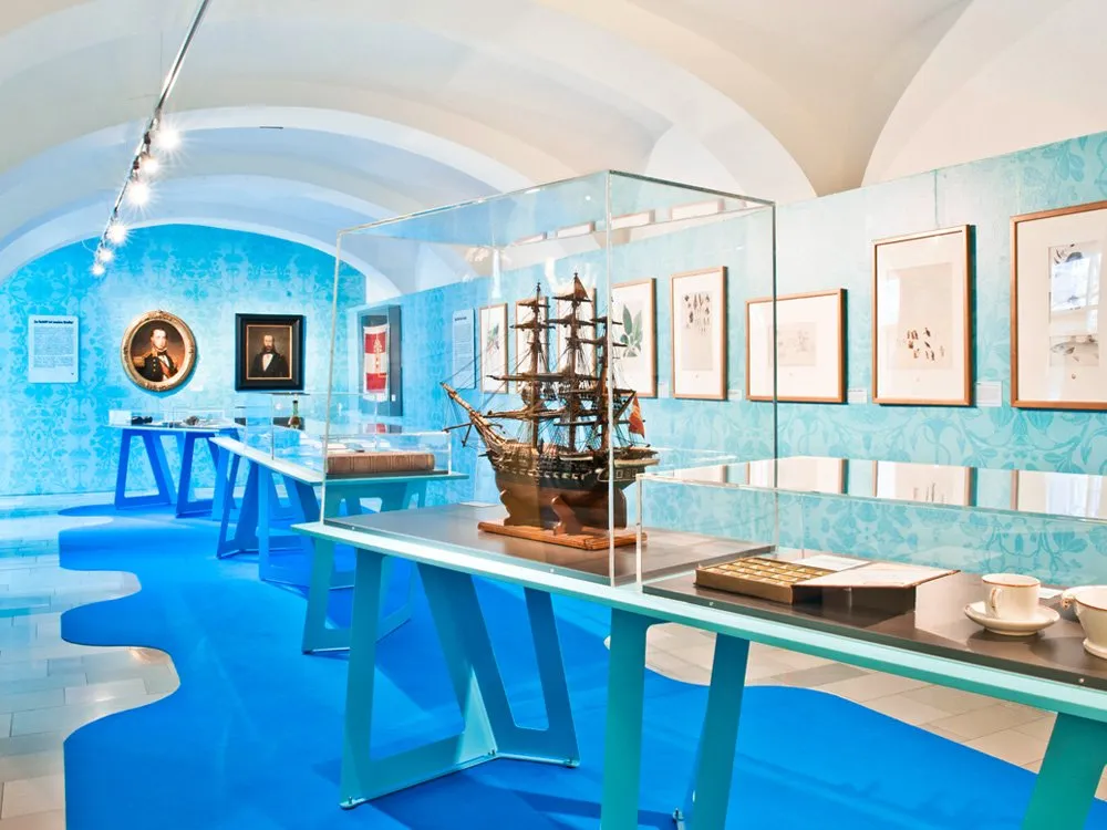 Ausstellungsraum: Blauer Raum mit gewölbter Decke, Vitrinen mit einem Schiffsmodell und Tassen, an den Wänden hängen Bilder und Porträts.