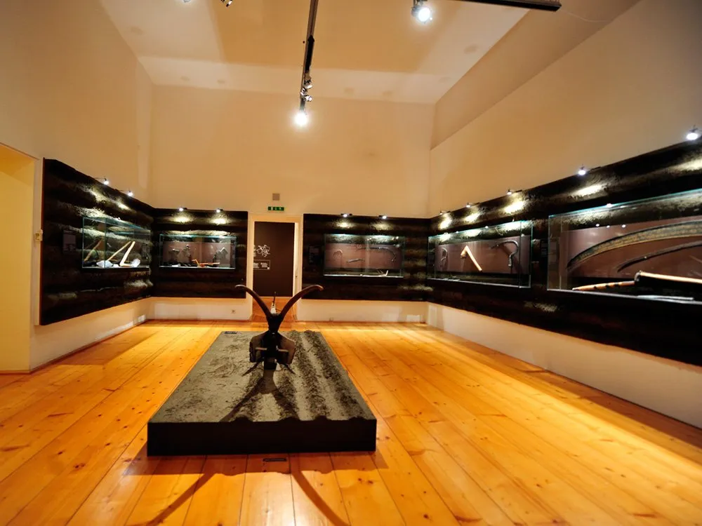 Ausstellungsraum: Helles Zimmer mit Holzboden, Vitrinen an den Wänden mit Ausstellungsstücken, zentrale Skulptur auf einem Podest