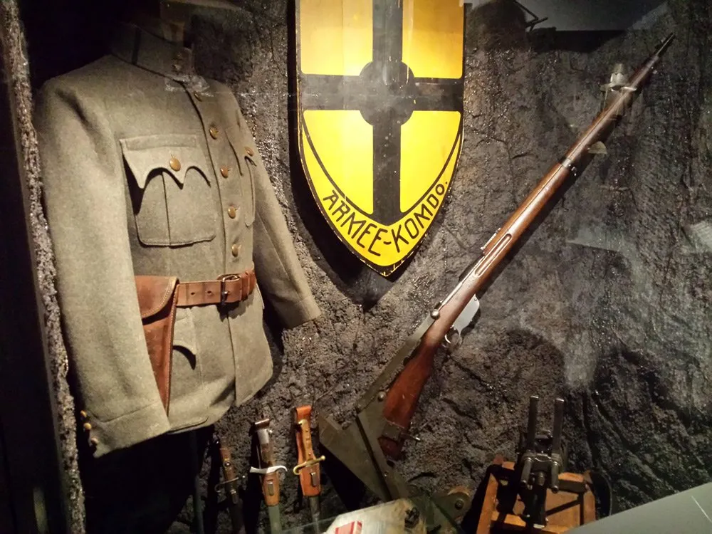 Ausstellungsraum: Eine Vitrine mit einer Militäruniform, einem Gewehr, Messern und einem Schild mit der Aufschrift "ARMEE-KOMDO" an einer steinernen Wand
