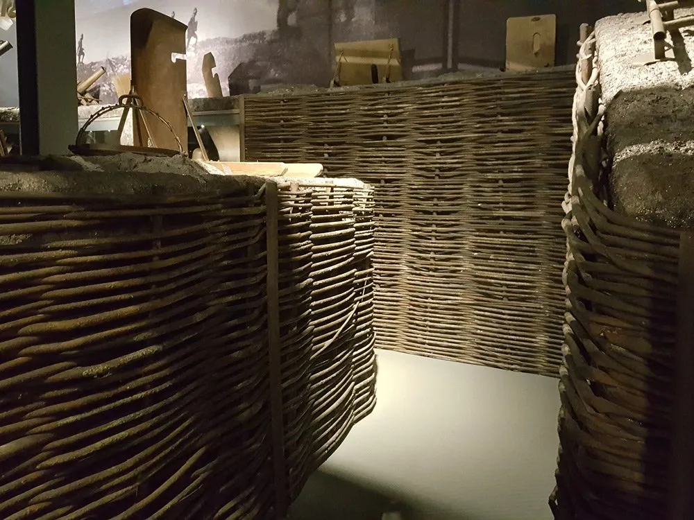 Ausstellungsraum: Eine Nachbildung eines Schützengrabens mit geflochtenen Wänden und historischen Objekten, darunter ein Schild und Stacheldraht, vor einem Bild von Soldaten