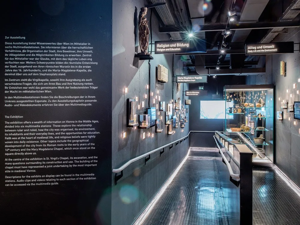 Ausstellungsraum: Dunkler Gang mit Vitrinen voller Artefakte, Schildern wie "Religion und Bildung" und "Alltag und Umwelt", sowie einer Statue und Multimedia-Bildschirmen an den Wänden.