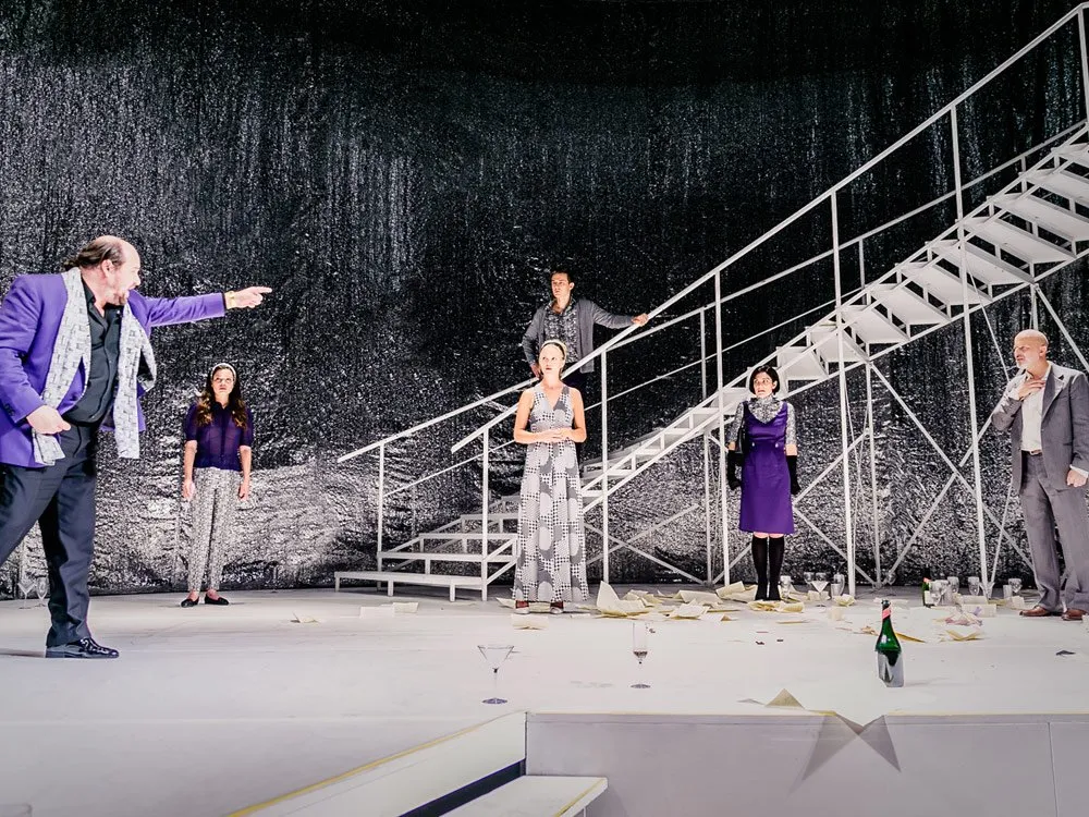 Szene: Theaterbühne mit weißer Treppe und glitzerndem Hintergrund. Fünf Schauspieler, einer in lila Anzug zeigt dramatisch. Gläser, Champagnerflasche und Papiere liegen verstreut.
