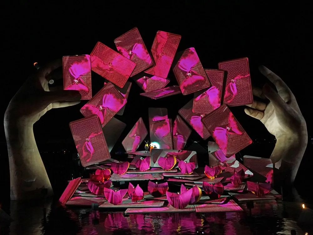 Szene: Große Hände halten eine Anordnung von rosa Spielkarten, Tänzer in bunten Kostümen