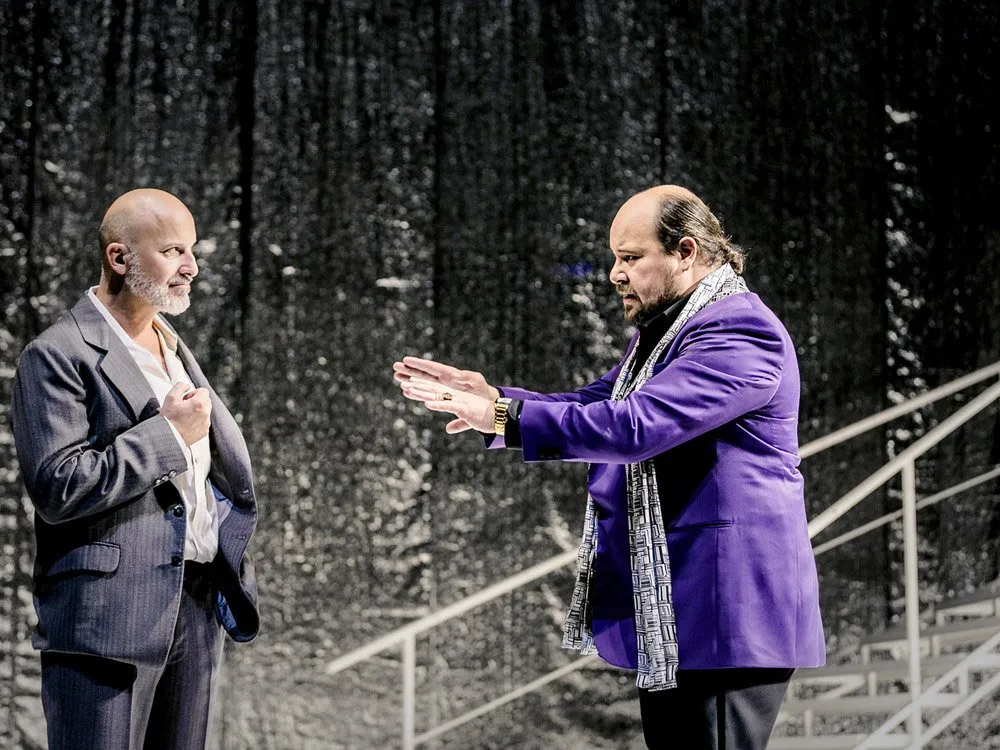 Szene: Zwei Schauspieler auf einer Bühne mit glitzerndem Hintergrund und weißer Treppe. Einer trägt einen grauen Anzug, der andere einen lila Anzug mit Schal und gestikuliert lebhaft.