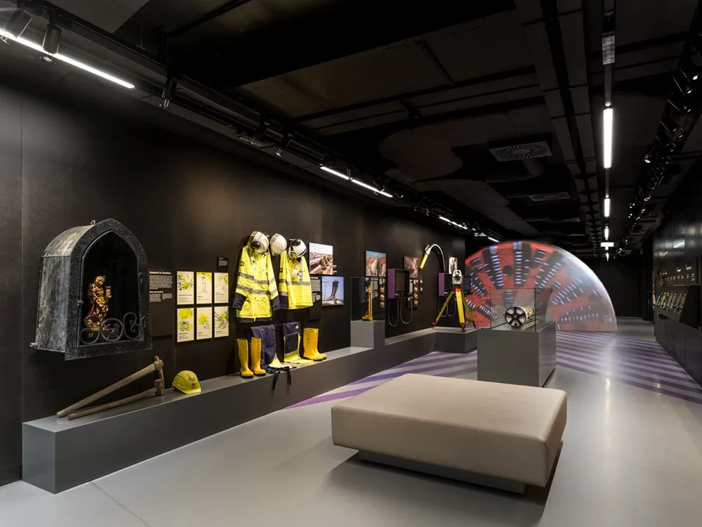 Ausstellungsraum: Dunkler Hintergrund, gelbe Schutzanzüge, Stiefel, Helm, Werkzeug, historische Nische mit Figur, interaktives Display, Theodolit, Sitzbank