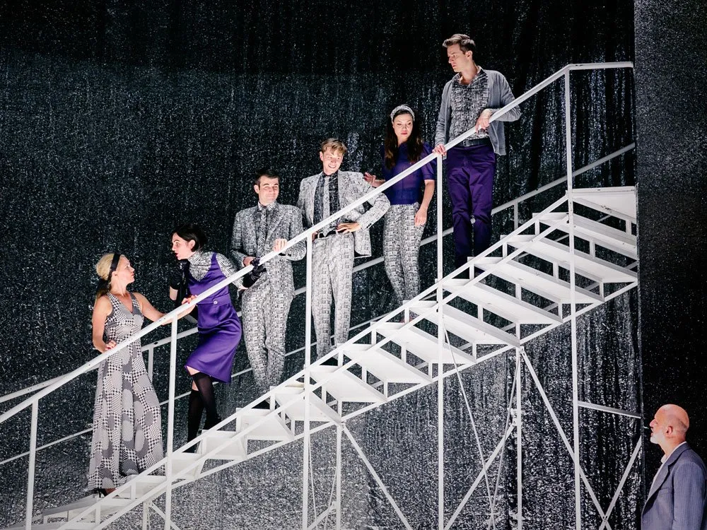 Szene: Sechs Schauspieler auf einer weißen Treppe mit glitzerndem Hintergrund. Drei tragen graue Anzüge mit Mustern, zwei sind in lila gekleidet, einer in einem grauen Kleid. Ein Mann in einem grauen Anzug steht unten rechts.
