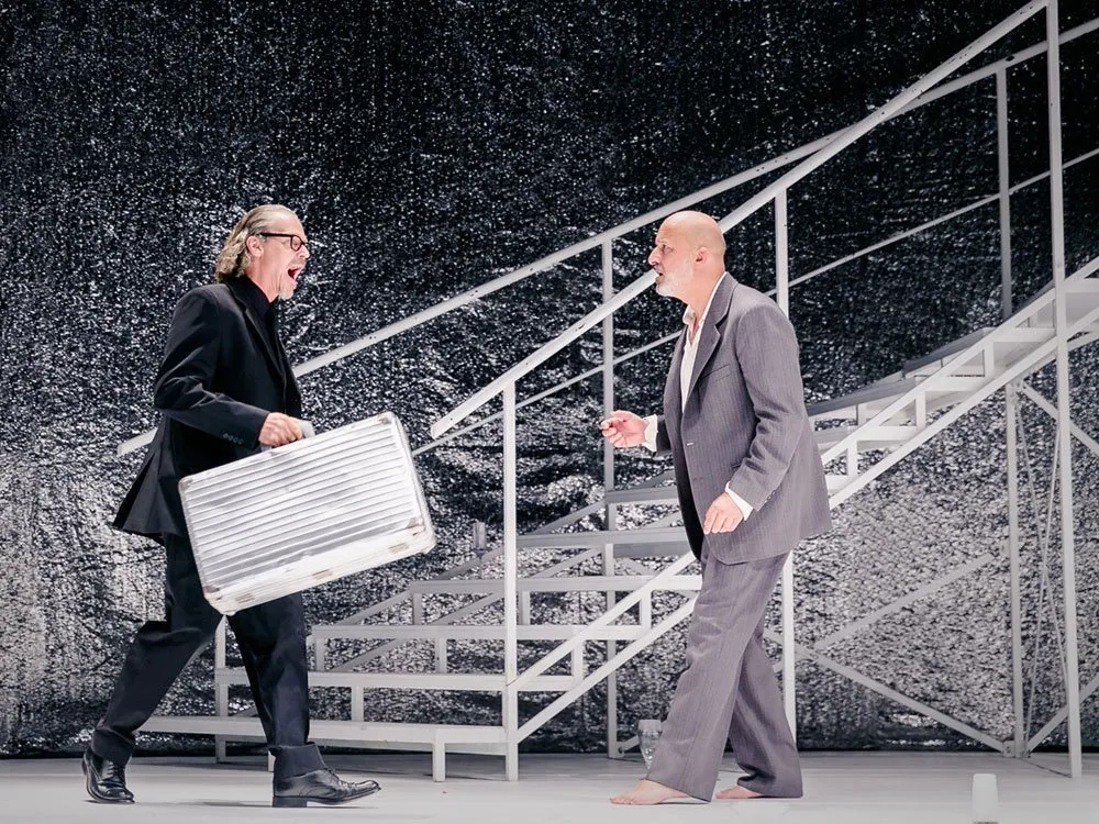 Szene: Zwei Schauspieler auf einer Bühne mit glitzerndem Hintergrund und weißer Treppe. Einer trägt einen schwarzen Anzug, hält einen Koffer und gestikuliert lebhaft. Der andere, barfuß in grauem Anzug, steht ruhig daneben.