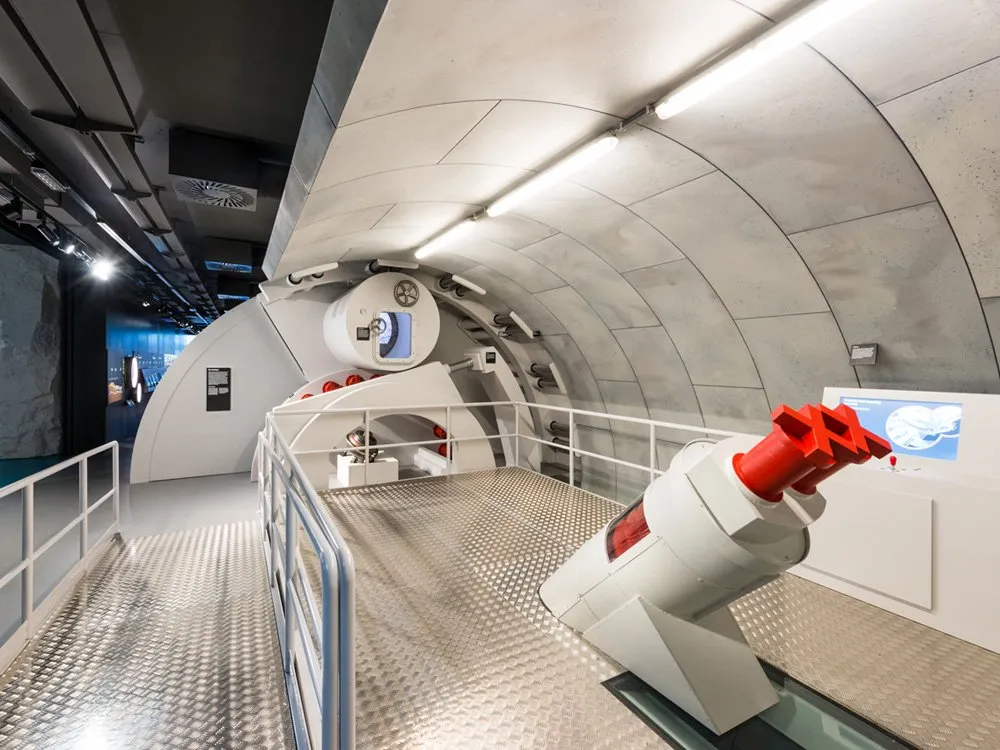 Ausstellungsraum: Eine Plattform mit Metallboden und Geländern, eine weiße Struktur mit rotem Element und ein rundes Modul mit Fenster