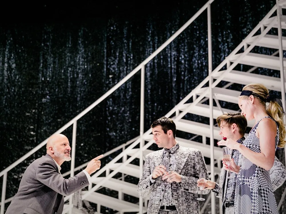 Szene: Ein Mann in grauem Anzug zeigt auf eine weiße Treppe mit glitzerndem Hintergrund. Drei Personen in gemusterten Outfits, darunter ein Mann in einem grauen Anzug mit Krawatte und zwei Frauen mit Champagnergläsern, stehen daneben.