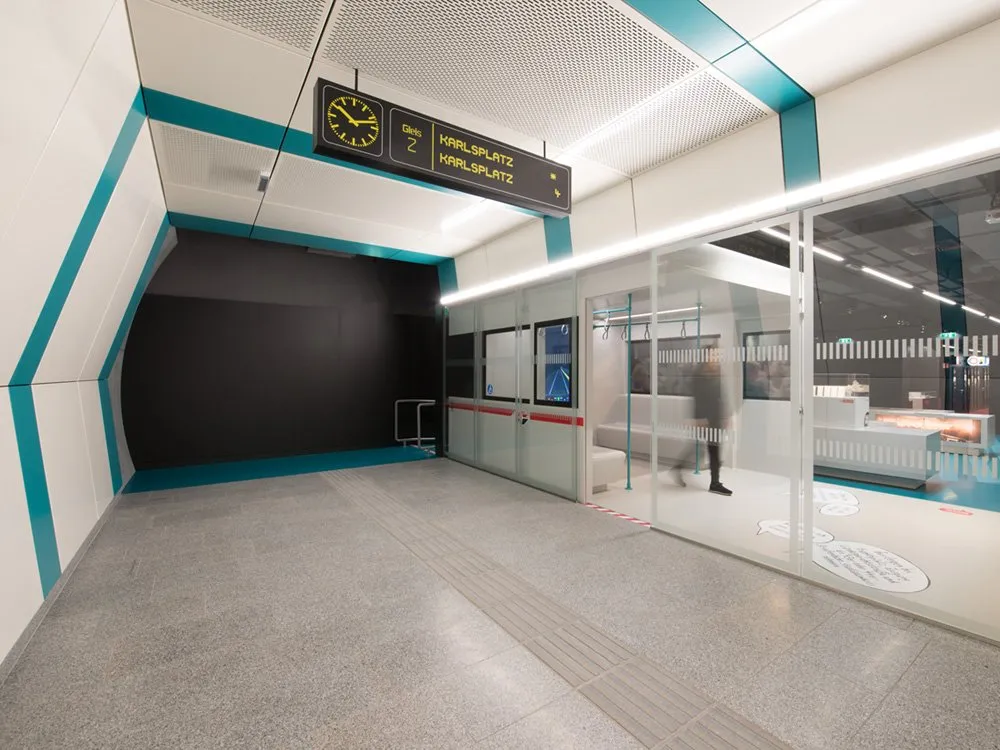 Ausstellungsraum: Moderne U-Bahn-Station mit Schild "Karlplatz", Uhr, Glastüren und einem Wartenden