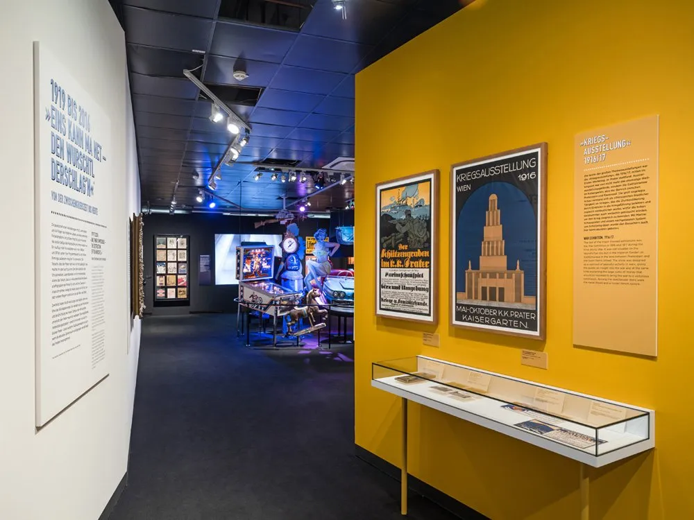 Ausstellungsraum: Historische Poster an gelber Wand, Ausstellungsstücke mit Karussellpferd und Auto