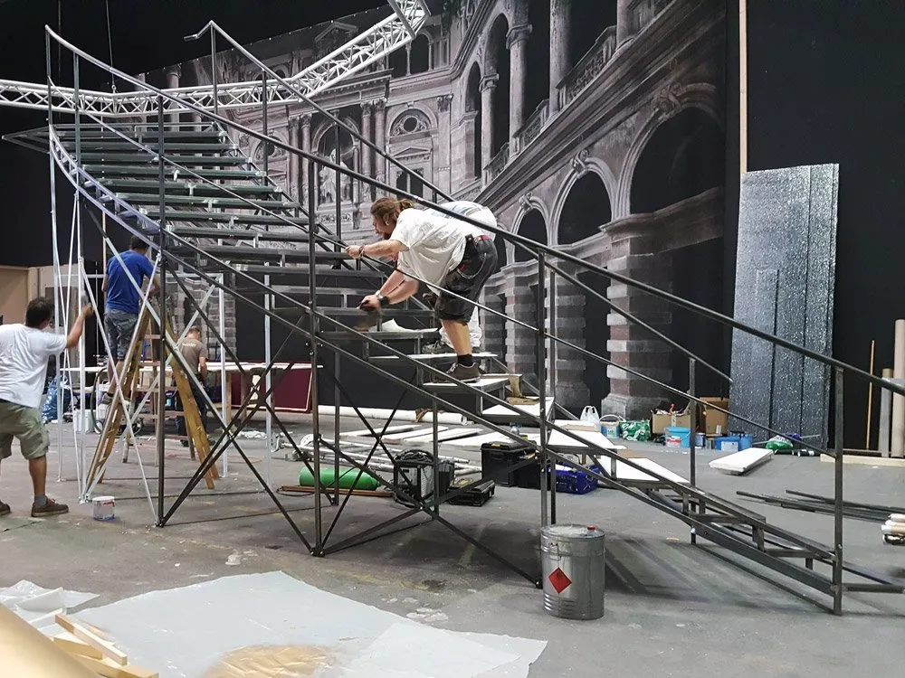 Szene: Eine Bühne im Bau mit einer schwarzen Metalltreppe vor einem Hintergrund mit klassischer Architektur. Drei Arbeiter sind aktiv, einer klettert die Treppe hoch, zwei andere arbeiten mit Werkzeugen und Leitern.