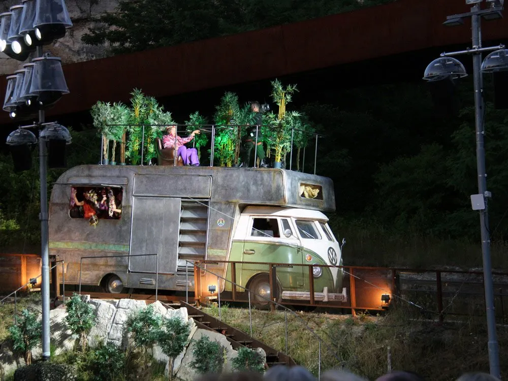 Szene: Verziertes VW-Bus-Modell mit Pflanzen auf dem Dach auf einer Bühne bei Nacht. Person in lila sitzt oben, andere schauen heraus