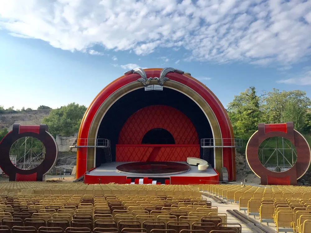 Szene: Rote Freiluftbühne mit Drehscheibe und Zuschauerrängen, umgeben von runden Strukturen und grünen Hügeln unter blauem Himmel.