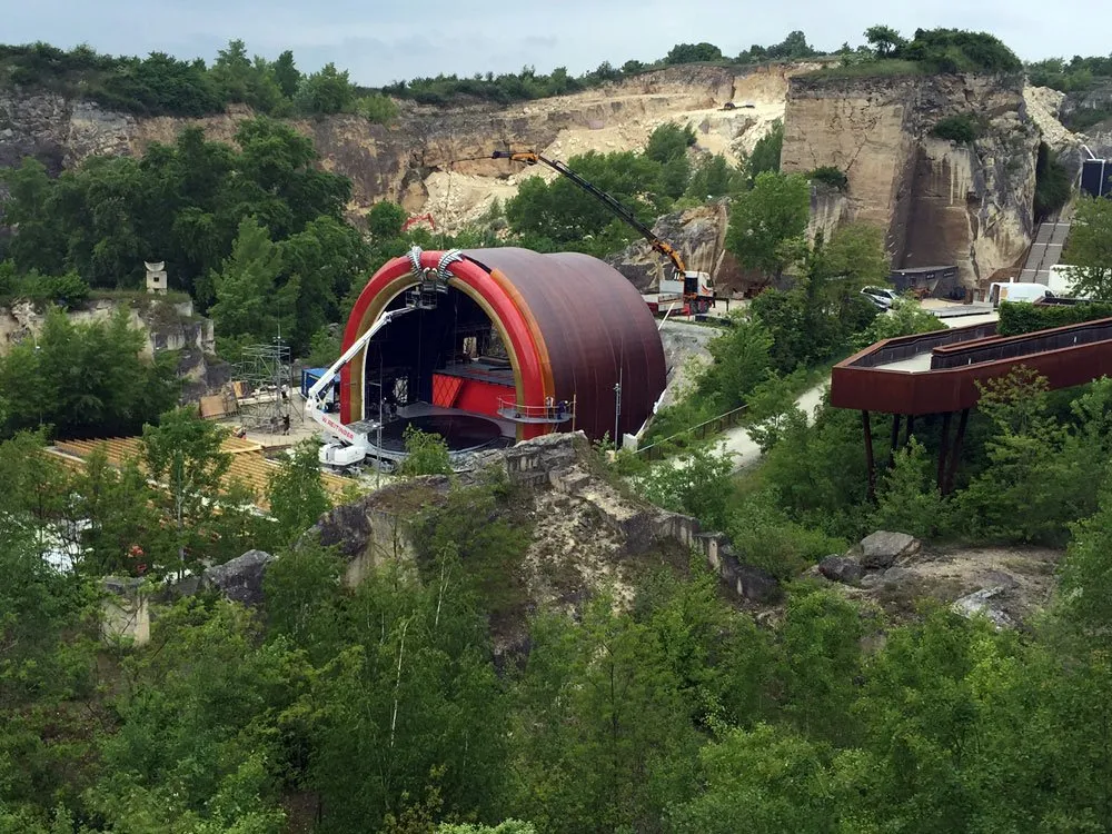 Szene: Rote Bühne in einem grünen, felsigen Tal mit Baumaschinen, Kran und Brücke im Hintergrund.