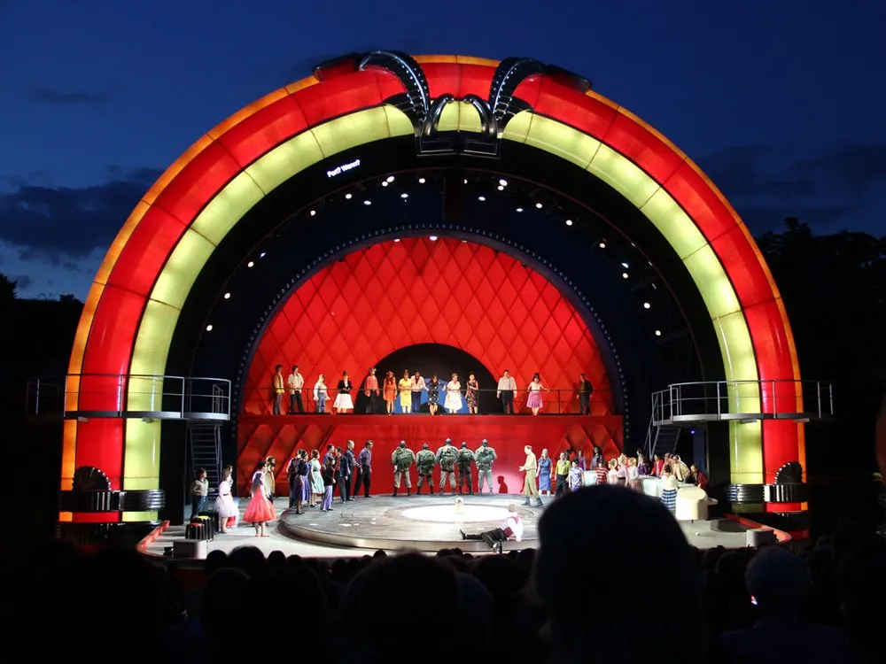 Szene: Bunte Freilichtbühne bei Nacht mit rot-gelber Rundung. Schauspieler in bunter Kleidung und Uniformen tanzen oder stehen auf einer Plattform, Publikum vor der Bühne