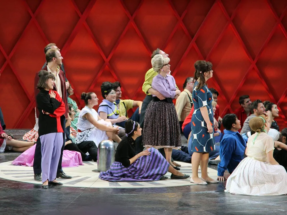 Szene: Schauspieler auf roter Bühne mit geometrischem Muster, in bunter retroartiger Kleidung, einige sitzen oder liegen, mit einem silbernen Zylinder.