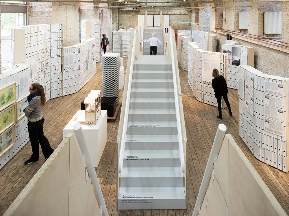 Ausstellungsraum mit Holzboden, weißen Wänden, Modellen und Dokumenten. Breite Treppe zentral im Raum, Besucher schauen sich um