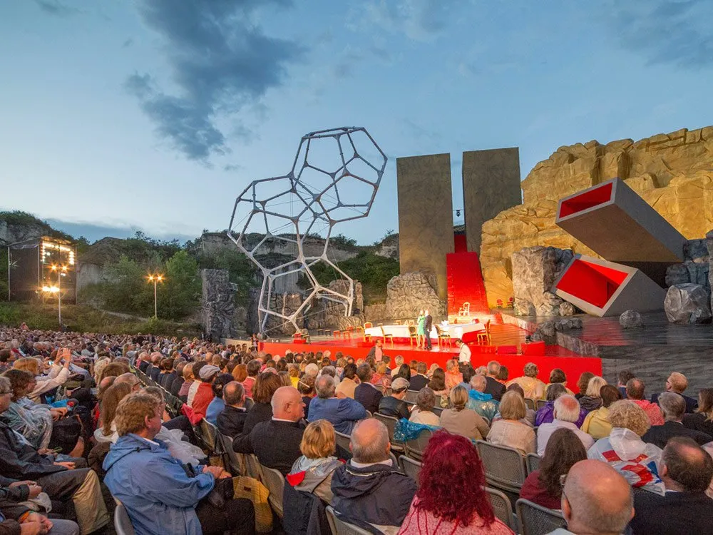 Theaterbühne mit roter Bühnenfläche, geometrischer Skulptur und Felskulissen, Publikum auf Sitzen, beleuchtet bei Dämmerung.