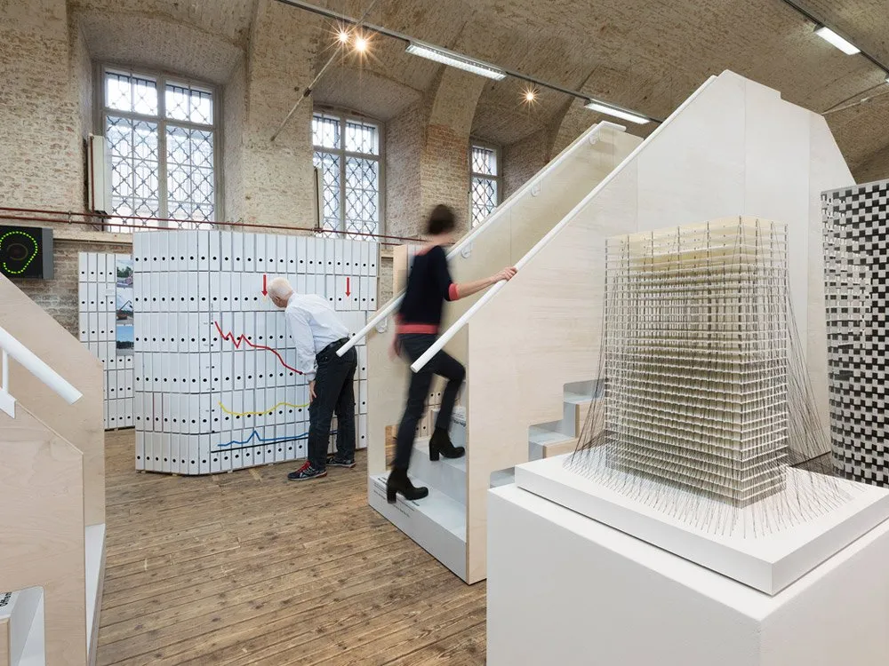 Ausstellungsraum mit Holzboden, Person auf Treppe, andere vor Diagrammen, Gebäudemodell auf Podest