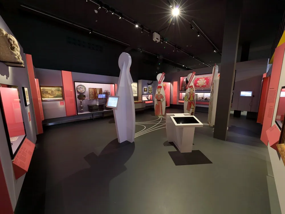 Ausstellungsraum: Moderner Raum mit rosa Wänden, Kunstwerken, Bildschirmen und stilisierten Figuren, beleuchtet von Deckenstrahlern