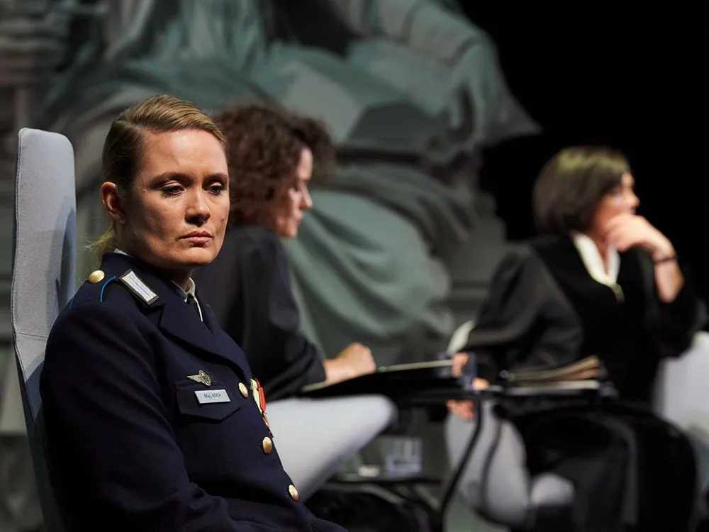 Szene: Uniform mit Namensschild "Maj Koch" sitzt auf einem Stuhl, umgeben von weiteren Personen in Roben, vor einer großen Statue im Hintergrund