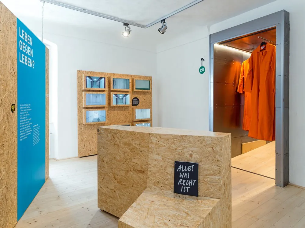 Ausstellungsraum: Helles Zimmer mit blauen und Holzwänden, einer Aufschrift "LEBEN GEGEN LEBEN?", einem Schild "ALLES WAS RECHT IST" und orangefarbenen Gefängnisanzügen.