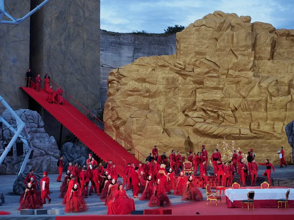 Szene: Rote Plattform und Treppe, goldene Felskulissen, viele Darsteller in roten Kostümen mit Masken, beleuchtet bei Dämmerung.