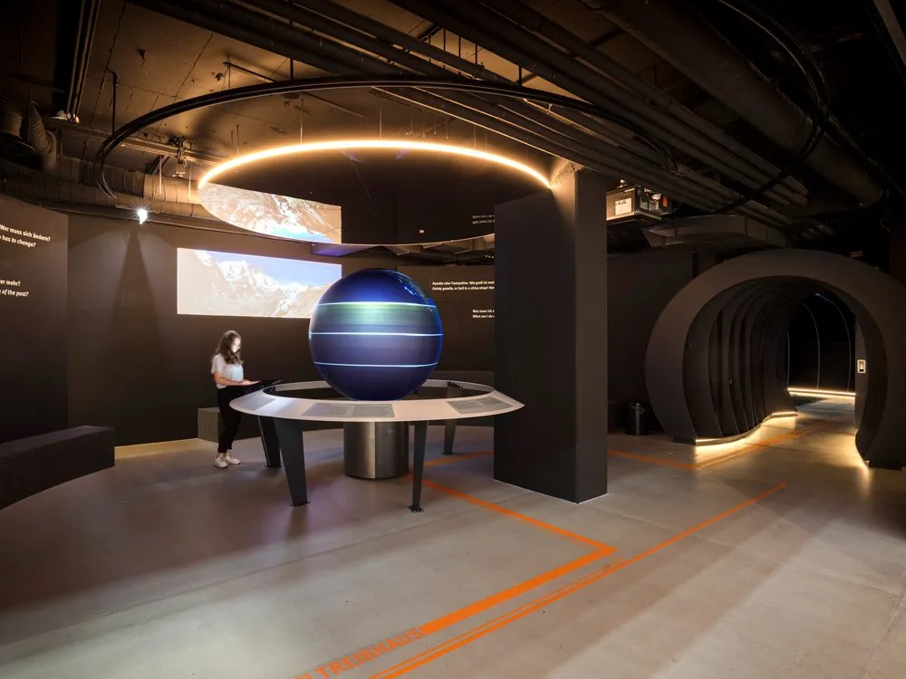 Ausstellungsraum: Person betrachtet eine leuchtende, blaue Erdkugel auf einem Tisch, mit einer Bergprojektion an der Wand