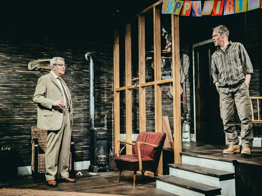Szene: Zwei Männer in einem rustikalen Raum mit Holzverkleidung, einer steht im Anzug vor einem Korbstuhl, der andere auf einer Treppe mit "Happy Birthday" Girlande, roter Sessel und Ofen im Hintergrund