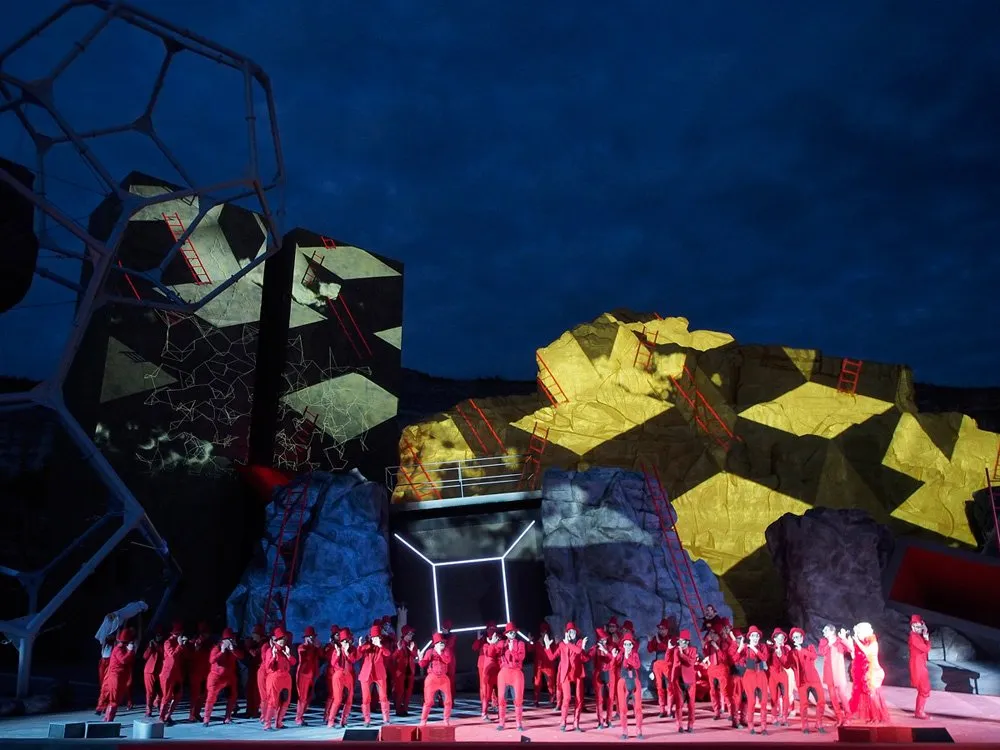 Szene: Viele Darsteller in roten Kostümen, geometrische und felsige Kulissen, beleuchtet bei Nacht mit blauen und gelben Lichtern.