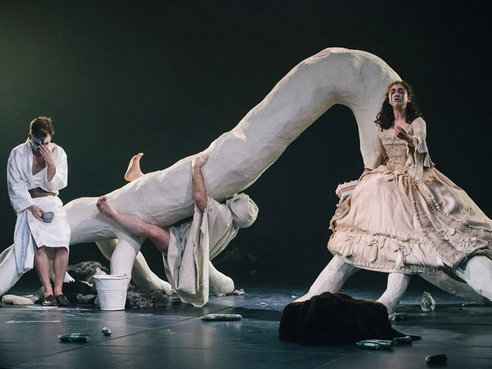 Szene: Drei Personen auf einer Bühne, eine sitzt in historischer Kleidung auf einer weißen Skulptur, eine klettert darauf, eine steht mit Eimer und Tasse, umgeben von Gegenständen auf dem Boden