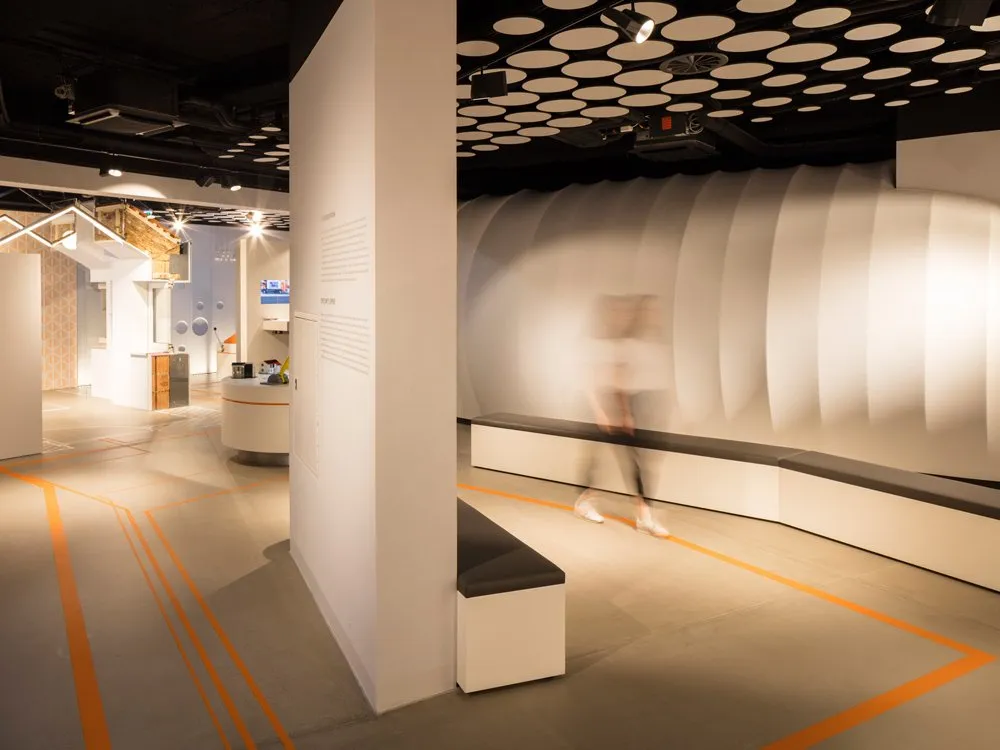 Ausstellungsraum: Weiße Wand mit Text, gewölbte Struktur, Deckenlampen, orangefarbene Linien am Boden