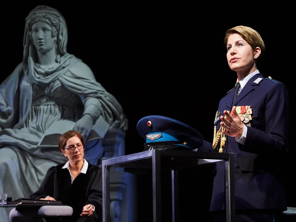 Szene: Person in Uniform mit Medaillen am Podium, eine in Robe sitzend, Justitia-Statue im Hintergrund.