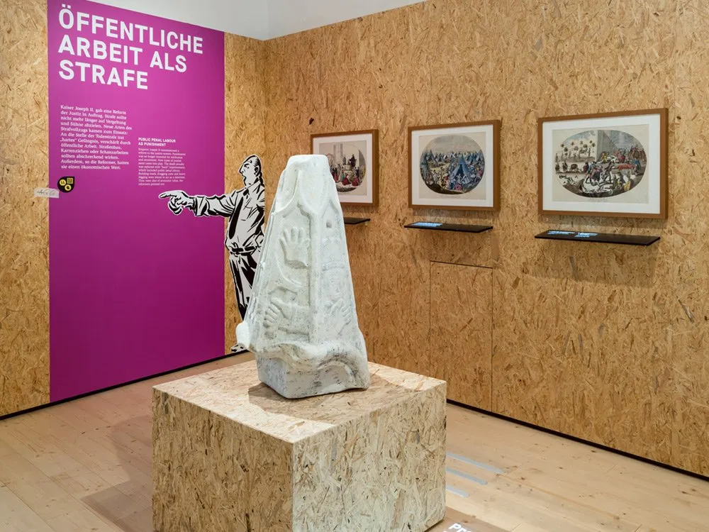 Ausstellungsraum: Lila und Holzstruktur mit der Aufschrift "ÖFFENTLICHE ARBEIT ALS STRAFE", einer Skulptur und historischen Bildern an der Wand.