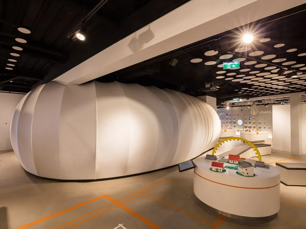 Ausstellungsraum: gewölbte, weiße Struktur, ein runder Tisch mit Modellhäusern und einem gelben Bogen