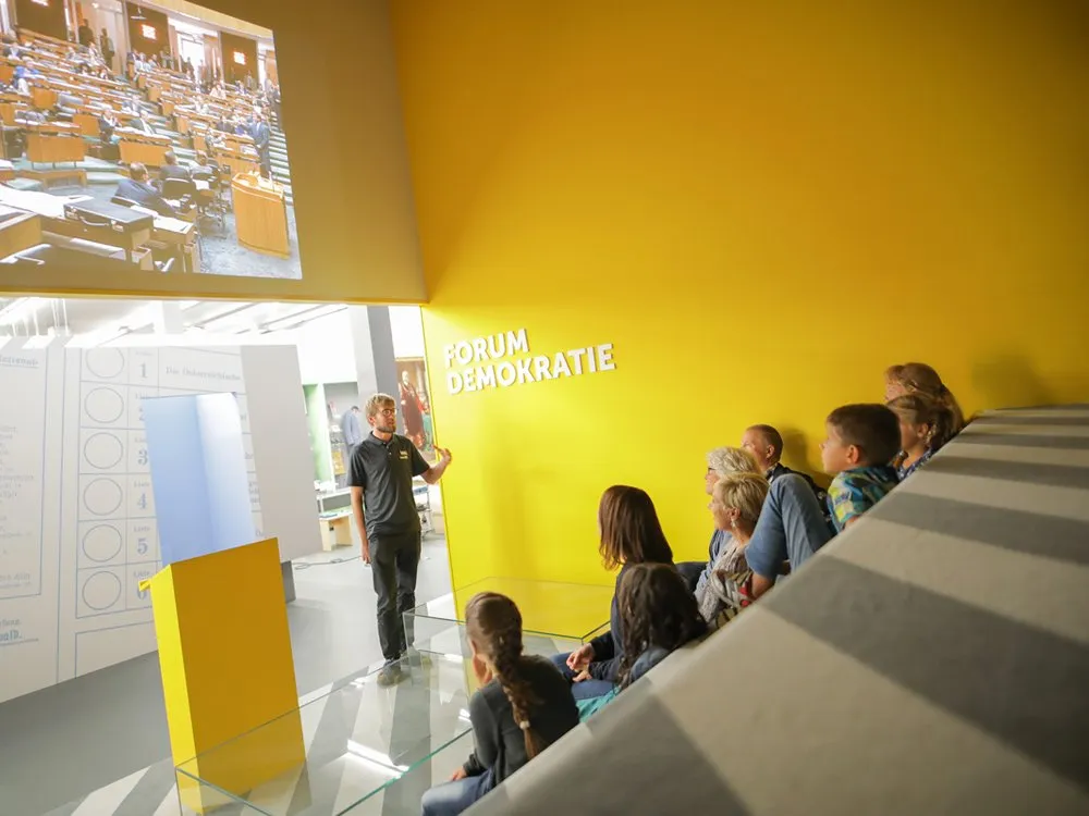 Ausstellungsraum: Ein Mann erklärt einer Gruppe Kinder vor einer gelben Wand mit der Aufschrift "FORUM DEMOKRATIE", während ein Bild eines Parlaments gezeigt wird.