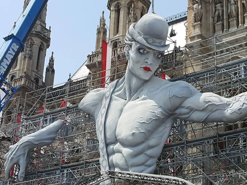 Große Statue eines Mannes vor dem Wiener Rathaus, umgeben von Gerüst und Kran, roter und weißer Hintergrund, sonniger Tag
