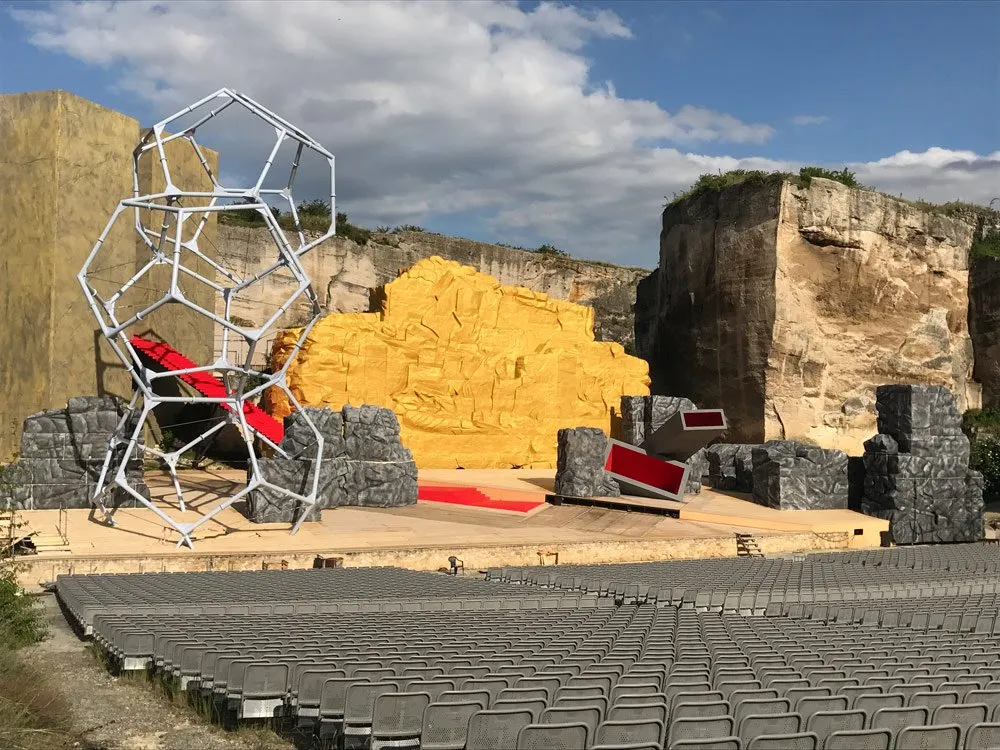 Szene: Geometrische weiße Skulptur, gelbe und graue Felskulissen, rote Elemente, leere Sitzreihen 