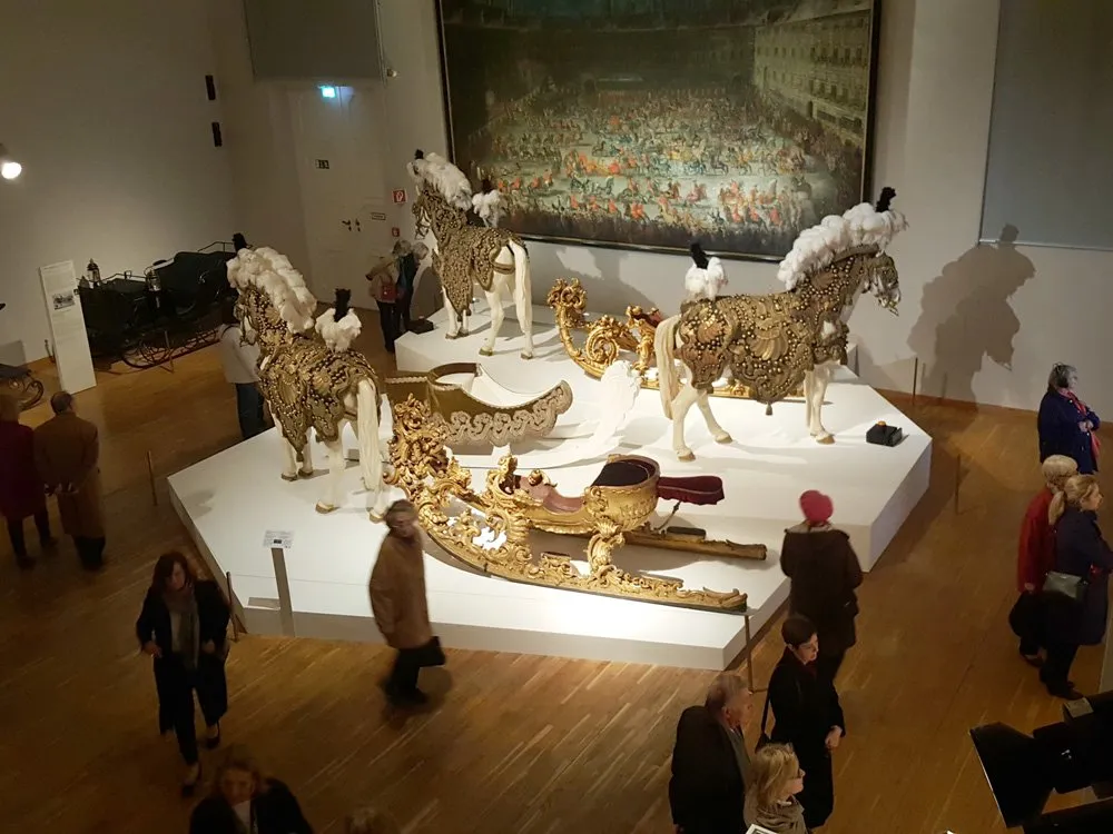 Ausstellungsraum: Goldene Kutsche mit Pferdefiguren auf Plattform, großes Gemälde an der Wand, Besucher umherlaufend