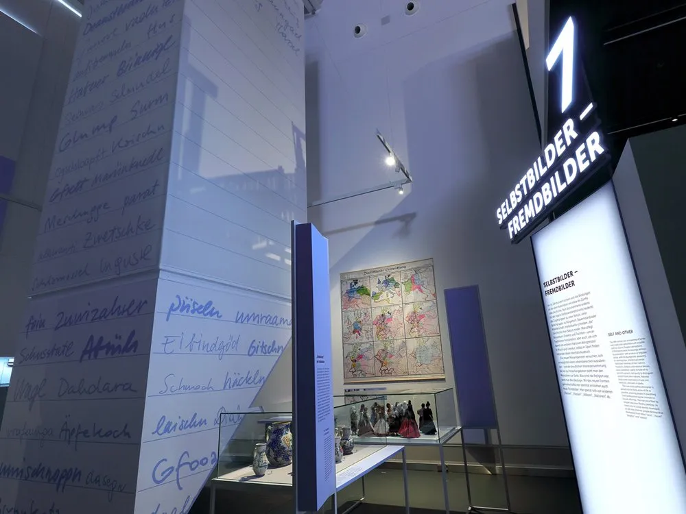 Ausstellungsraum: Eine Wand mit handschriftlichen Notizen, eine Karte und eine Vitrine mit Figuren unter dem Thema "Selbstbilder – Fremdbilder"