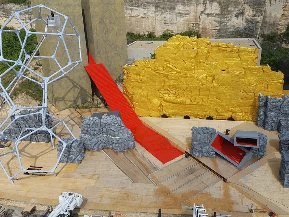 Szene: Geometrische weiße Skulptur, gelbe und graue Felskulissen, rote Treppe, Baukran und Arbeiter bei der Vorbereitung, heller Tageshimmel