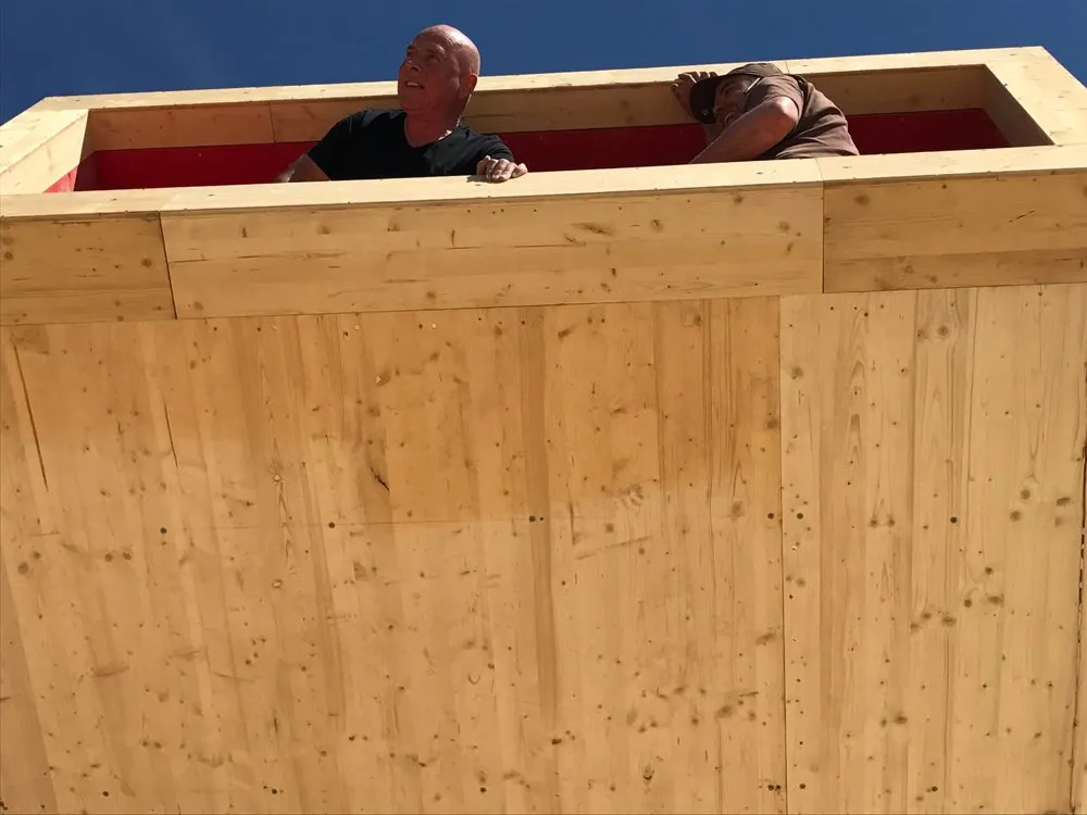 Szene: Zwei Arbeiter in einer großen hölzernen Konstruktion mit roter Innenwand, heller Tageshimmel im Hintergrund