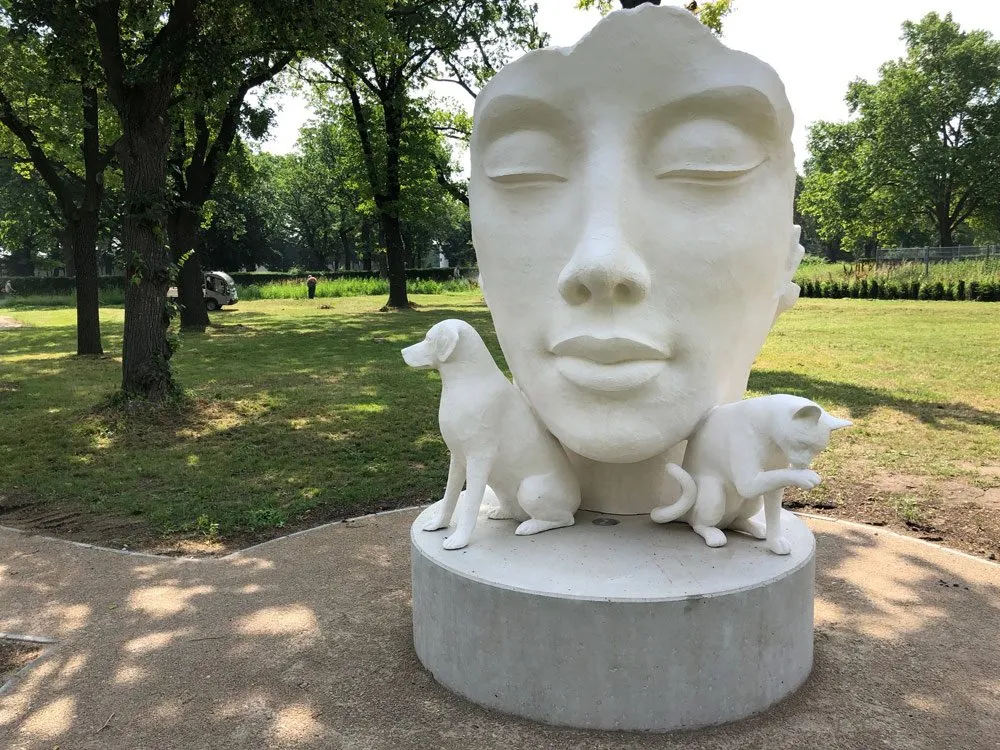 Weiße Skulptur mit Gesicht, Hund und Katze auf einem Sockel in einem grünen Park