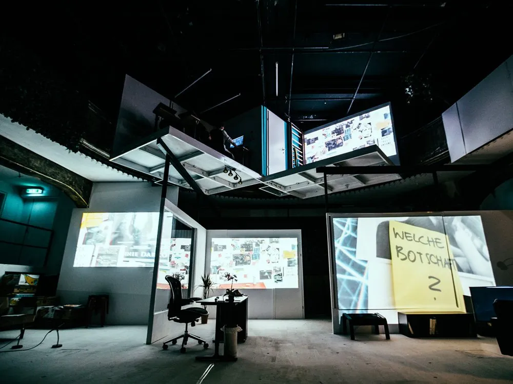 Theaterraum mit Leinwänden, darunter "Welche Botschaft?", einem Schreibtisch, Stuhl und Pflanzen im dunklen Ambiente