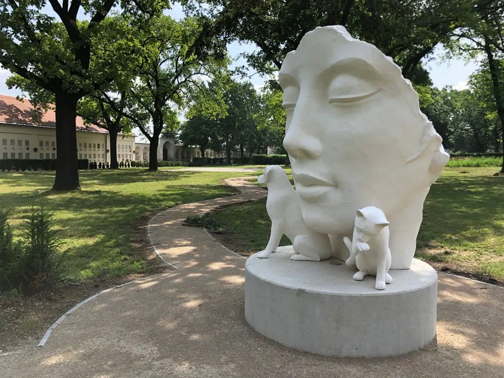 Weiße Skulptur mit Gesicht, Hund und Katze auf einem Sockel in einem Park mit Bäumen und einem Gebäude im Hintergrund