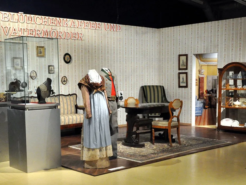Ein Raum mit gestreifter Tapete zeigt ein Kleid und Uniformrock. Vitrine mit Fotos, Sofa, Tisch, Stühle und Schrank mit Porzellan. Text "BLÜMCHENKAFFEE UND VATERMÖRDER" an der Wand.