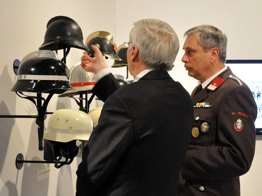 Zwei Personen betrachten eine Reihe historischer Helme, darunter schwarze, goldene und weiße Modelle, an einer Wand ausgestellt