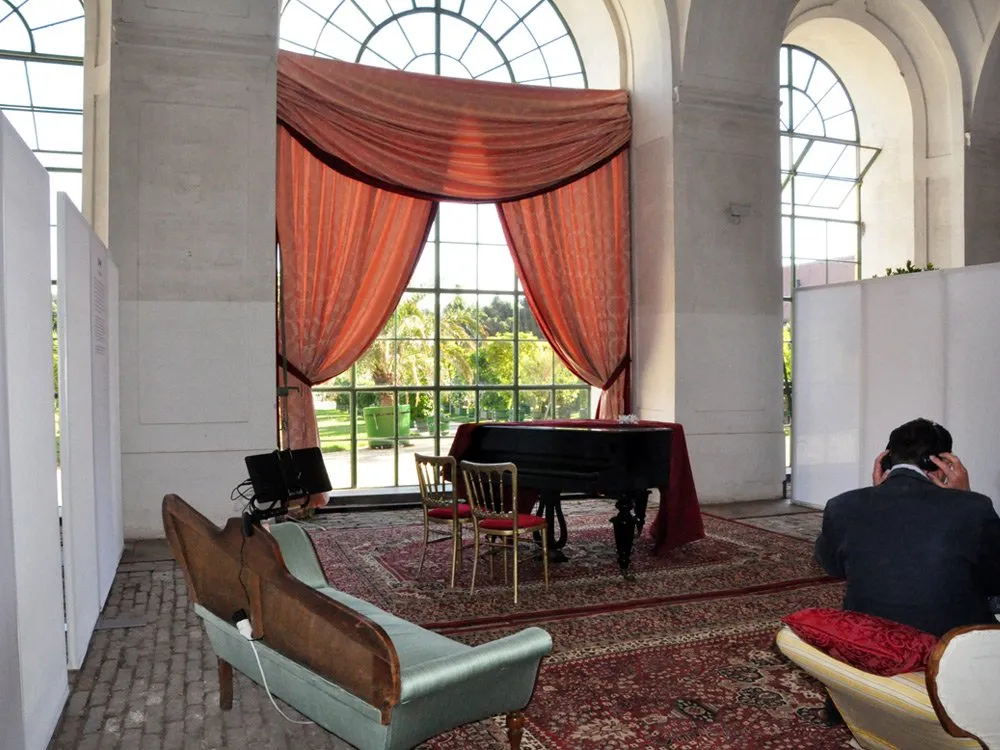 Ausstellungsraum mit einem Flügel unter großen Fenstern mit orangefarbenen Vorhängen, umgeben von antiken Möbeln und einem orientalischen Teppich